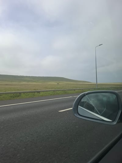 An overcast sky hangs above a vast, rolling landscape of green and brown fields, bordered by a distant hill. A road stretches into the distance, marked with a white line, with a metal guardrail separating it from the fields. A single streetlamp stands tall against the horizon. In the foreground, a car's side mirror reflects the scene, offering a distorted view of the road and the sky. The image appears to be taken from inside a moving vehicle, capturing the essence of a journey through a rural, undeveloped area. The location is not identified as being in Calderdale, United Kingdom.