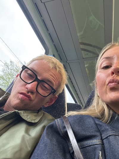 A selfie taken from below features two individuals. The person in the lower half of the frame is a man with light blond hair, wearing black-framed glasses and a jacket. His eyes are partially closed and he appears to be looking down. Above him is a woman with blond hair, pouting towards the camera. She is wearing a dark denim jacket. The background suggests they are on a train. The location is Welwyn Hatfield, United Kingdom.