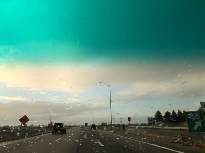 A first-person perspective shot from inside a vehicle traveling on a highway. The windshield is speckled with raindrops. The sky is overcast with hues of turquoise, beige, and grey. Several cars are visible on the road. On the left side of the highway, there is a construction zone with an orange diamond-shaped sign featuring a silhouette of a falling person. Further down the road, on the right, a green sign indicates "35" with an arrow pointing right, suggesting an exit. In the background, there are trees and buildings, including a sign that appears to read "Royal Homes." The scene suggests a drive through Innisfil, Canada, possibly during a light rain shower.
