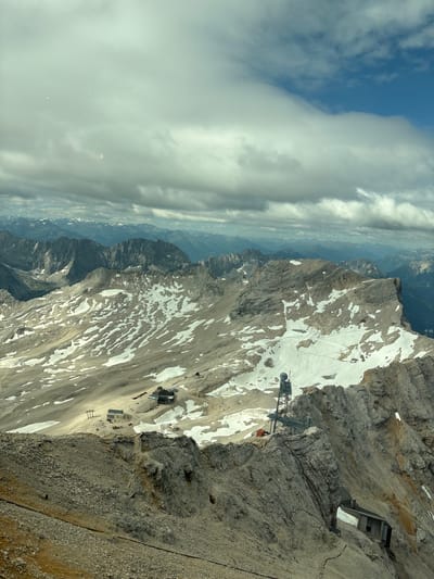 This image is a panoramic view of a mountainous landscape, likely taken from a high vantage point. The foreground features a rocky, uneven terrain with sparse vegetation. In the mid-ground, a vast mountain range unfolds, characterized by steep, rugged peaks and valleys. Patches of snow are visible on the slopes, indicating a high altitude. Several man-made structures are present, including buildings, ski lifts, and a prominent radio tower with antennas, all appearing to be built into or on the mountainside. The background reveals a layered landscape of more distant mountains fading into a hazy horizon under a cloudy sky with patches of blue. The lighting suggests it is daytime. While the prompt mentions Ehrwald, Austria, the visual elements are consistent with a high alpine environment. There are no individuals or discernible actions or interactions depicted in the image.