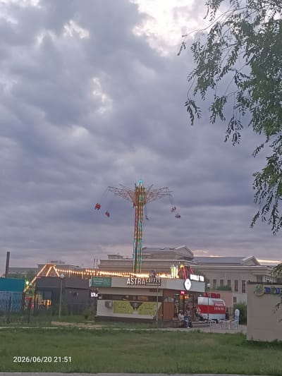 The image shows an amusement park ride with people on it against a cloudy sky. In the foreground, there is a cafe with "ASTRA COFFEE" and "BUBBLE TEA" signage. A red and white food truck is parked nearby. Several people are sitting at tables outside the cafe. In the background, there are buildings. A timestamp at the bottom left corner indicates "2026/06/20 21:51". The scene is likely in the city of Oral, Kazakhstan, given the style of the cafe and food truck, and the Cyrillic script on the signage.