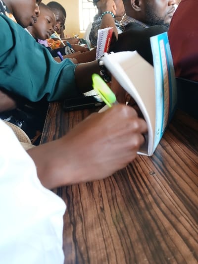 The scene is set indoors in Ugi Majowel, Nigeria, depicting an educational or training environment with multiple individuals seated at wooden desks. In the immediate foreground, a dark-skinned hand, partially covered by a white sleeve, actively writes in an open white spiral-bound notebook with a blue spine, using a neon green pen. A dark-colored smartphone rests on the dark-grained wooden desk to the left of the writing hand.
In the mid-ground, from left to right, a dark-skinned individual wearing a teal long-sleeved shirt is visible from the chest up, facing right. Adjacent to them, another younger dark-skinned individual is seated, looking forward. Further to the right, a dark-skinned individual with a beard, wearing a patterned shirt, is partially visible. Another person wears a beaded bracelet on their wrist and holds a red spiral notebook.
The background consists of a light-colored wall, with a window featuring security bars visible in the upper portion, allowing natural light into the space. The individuals appear attentive, seated, and engaged in activities consistent with a classroom or lecture setting, primarily note-taking and observation.