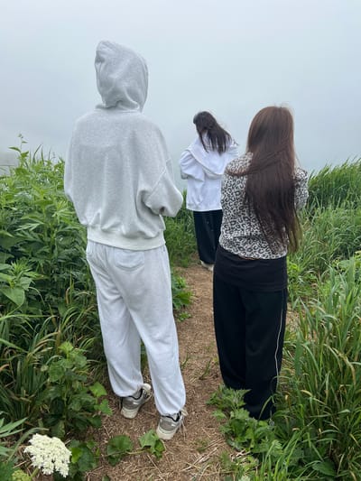 Three individuals are standing on a dirt path in a grassy area with a faint white fog or mist in the background. The person in the foreground is wearing a grey hooded sweatshirt and grey sweatpants, with their back to the camera. To their right and slightly behind them, another person is wearing a white hooded sweatshirt and black pants, also with their back to the camera. Further back and to the right, a third person is wearing a leopard-print sweatshirt and black pants, facing away from the camera. The ground is covered in dry grass and dirt. The vegetation around the path is lush and green. The location is not identifiable as Tsapko, Russia from the image content.
