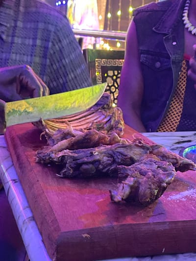 An indoor, close-up shot captures a person in Kimorori, Kenya, preparing to eat a meal. The scene is bathed in dim, colorful artificial lighting, dominated by purple and blue tones, indicating an evening or nighttime setting.
In the foreground and central-right, a large portion of cooked meat, resembling ribs or bone-in cuts, rests on a reddish-brown wooden cutting board. A light green or yellowish-bladed knife with a dark handle is held by a hand in the lower-left, positioned as if ready to carve the meat. White residue, possibly salt or spices, is scattered on the board near the food.
Partially visible on the left is the upper torso of a person, wearing a dark denim vest over a patterned garment. A white beaded necklace is visible around their neck. The background features blurry lights and a dark surface with a gold Greek key pattern, suggesting the decor of a restaurant or eatery. No discernible text is present in the image.