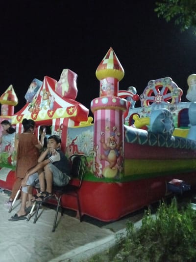 In Dustlik, Uzbekistan, at night, a large, colorful inflatable circus-themed play structure is illuminated. The structure features red and white striped tents, towers with red and yellow checkered tops, and various cartoonish characters like a bear juggling balls and other circus performers. In the foreground, two young boys are seated on a metal chair placed in front of the play structure. One boy, wearing a dark shirt with white trim and light blue shorts, is sitting with his arms crossed. The other boy, also wearing a dark shirt and light shorts, is partially obscured by the first boy and leans back in his seat. A woman in a brown tunic and pants stands behind the boys, facing away from the camera and holding a smartphone. Her left hand is raised, possibly operating the phone. The ground beneath them appears to be paved with large, light-colored stone tiles, and dark green foliage is visible to the right and in the background, indicating an outdoor setting.