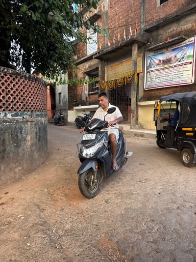 This image captures an outdoor street scene in Gokarna, Karnataka, India. The location is confirmed by the "KA 47" RTO code on the scooter's license plate (Karwar district, which includes Gokarna) and Kannada script banners on a building, one of which mentions "ಮಹಾಬಲೇಶ್ವರ ದೇವಸ್ಥಾನ" (Mahabaleshwar Temple), a prominent landmark in Gokarna.

A single adult male is the central figure, seated on a dark grey Honda Dio scooter bearing license plate "KA 47 Y 0992." The scooter's front cowl displays a red mark, possibly a tilak, and it is equipped with a chrome front crash guard. The man has short dark hair, a mustache, and a short beard. He wears a light-colored, short-sleeved collared shirt, light-colored shorts, and dark flip-flops. His hands rest on the handlebars, and his head faces forward.

The ground is an unpaved, dusty surface composed of dirt and gravel, exhibiting unevenness and scattered small stones. To the right of the scooter, a black and yellow auto-rickshaw is partially visible, appearing stationary.

In the background, a multi-story building constructed of reddish-brown brickwork and concrete features prominently. Its facade includes several windows, some fitted with security bars, and an upper window covered by a white sheet. The building's entrance is decorated with yellow and orange floral garlands, and a row of small, orange triangular flags is strung horizontally above the entrance. Two large banners are affixed to the building; the one on the right is white with images and text in Kannada script, while the left banner is yellow with black Kannada text. To the far left, a light brown wall with a circular lattice pattern and a tree with green leaves are partially visible, alongside a narrow alleyway where additional motorcycles are parked.