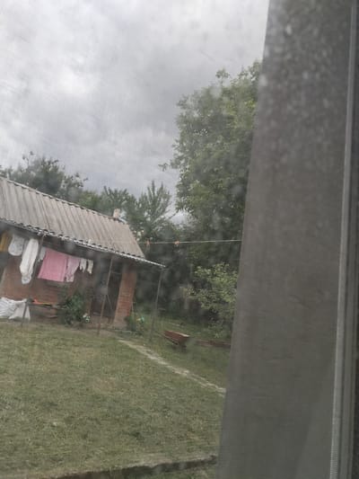 The image depicts a cloudy, overcast day in a rural setting, likely in Vysokyi, Ukraine. The foreground is dominated by a weathered, gray-tiled roof of a small brick house. Clothes hang on a clothesline strung across the front of the house, including a pink garment and several white items, suggesting laundry is being dried. A black dress with white polka dots is also visible hanging.
The house itself appears to be constructed of red brick and has a porch area. There are some white bags or sacks placed near the left side of the house. Green grass covers the yard, and a dirt path runs through it. In the background, several trees with dense green foliage are visible, their leaves appearing somewhat muted by the cloudy conditions.
To the right of the frame, a portion of a light gray, textured surface, possibly a window screen or a similar material, is present. The sky is a uniform shade of gray, indicating cloudy or potentially rainy weather. The overall atmosphere is subdued and somewhat somber, consistent with a gray day. There are no visible people or text in the image. The presence of laundry drying and the general appearance of the dwelling suggest a domestic, everyday scene.