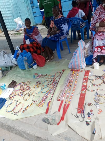 The image depicts an outdoor market stall in the Ildamat ward, Kenya. A light green tarp is spread on a paved surface, showcasing a collection of handcrafted Maasai-style beadwork. The merchandise includes numerous necklaces, bracelets, earrings, and several red leather belts, all intricately adorned with colorful geometric patterns in yellow, orange, green, blue, white, and black beads. Additionally, three pairs of flat shoes (two brown, one blue), two rolls of white paper, and a small clear plastic bottle are displayed on the tarp. A grey stone partially rests on the tarp's edge in the foreground.

Four individuals are positioned behind the display. To the immediate left, a person with short dark hair, wearing a blue long-sleeved shirt and a red, black, and white plaid garment, is seated on the ground, actively consuming an item held to their mouth. Adjacent to them, an older individual, likely female, with short dark hair and multiple necklaces, is seated on a blue plastic chair. They wear a patterned blue and white shawl over a light-colored top and a dark blue skirt, with light-colored flat shoes. Further right, two additional individuals are partially visible, seated on blue plastic chairs; one wears a red shawl over a striped top, and the other a white and red patterned garment.

In the background, a light blue corrugated metal wall or gate is present. Blurred figures are visible, including one person wearing a green t-shirt with the text "I TOUCHED WORLD'S HEART." Various bags, including blue, red, and white sacks, are placed near the seated individuals. A dark vertical pole is visible on the far left.
