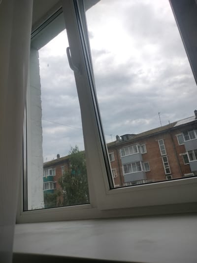 The image shows a view through a slightly open window, looking out onto a cloudy sky and a brick apartment building. The window frame is white plastic. On the left side of the frame, a white curtain is visible. A window handle is present on the right side of the window frame. The apartment building is made of red brick and has multiple windows and balconies. Some trees are visible in front of the building. The sky is overcast with gray clouds. The scene appears to be in a city in Russia, likely Osinniki, given the architectural style of the building.