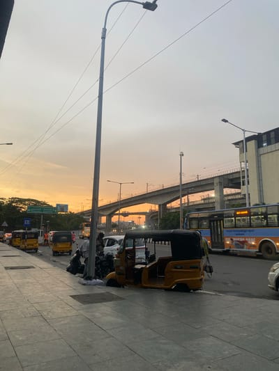 The image shows a street scene at dusk in St. Thomas Mount-Pallavaram Cantonment Board, India. The sky is a soft orange and gray, indicating sunset. A prominent elevated railway track or highway overpass dominates the middle ground, supported by concrete pillars. A blue and orange bus is visible on the right side of the road, and several yellow auto-rickshaws are present, some parked along the sidewalk and others in traffic. People are seen near the auto-rickshaws, some appearing to be drivers or passengers. The sidewalk is paved with gray tiles. A lamppost stands tall on the left side of the image, with wires extending from it. Traffic signs are visible in the distance, and trees line the far side of the road. The overall atmosphere is that of a busy urban environment during the transition from day to night.
