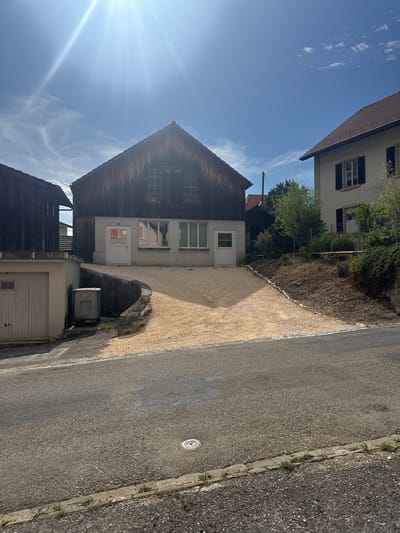 The image shows a sunny day in Court, Switzerland. In the foreground, there is a paved road with a concrete curb and a drain cover. Behind the curb, there is a light-colored gravel driveway leading up to a wooden building. This building has a dark-stained wooden facade and several windows. To the left of the wooden building, there is a lighter colored building with a garage door. To the right of the wooden building, there is a white building with a dark-tiled roof and windows. The sky is blue with some wispy clouds, and the sun is shining brightly, creating lens flare. The overall scene depicts a residential area with a mix of architectural styles.