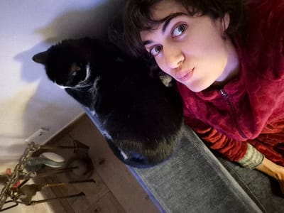 A person in a red hoodie poses for a selfie, with a black and white cat lying on their lap. The person is fair-skinned with short, dark hair and is looking directly at the camera with a slightly pursed mouth. The cat is positioned near the person's shoulder, facing away from the camera. The background features a light-colored wall and a decorative metal object with glass components. The person is wearing grey denim jeans.