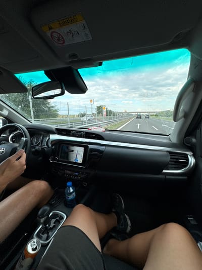 The image shows the interior of a car looking out towards a highway. The perspective is from the passenger seat, with a person's legs visible in the foreground. The car's dashboard is prominent, featuring a navigation screen displaying a map, a steering wheel, and various controls. Outside the car, a highway stretches into the distance with other vehicles on the road. There are signs indicating services like Shell and McDonald's, suggesting a location along a major transport route. The sky is cloudy. The image may have been taken in the vicinity of Radinovo, Bulgaria, as indicated by the prompt.