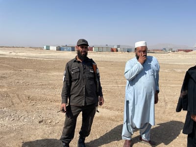 The image depicts an outdoor scene in Pir Dust, Afghanistan, under a clear, bright blue sky. The ground is an open, arid expanse of light brown dirt and gravel with visible tire tracks.
In the foreground and mid-ground, three individuals are present. On the left, a man with a dark beard and mustache, wearing a dark gray baseball cap, stands. He is dressed in a dark, long-sleeved uniform shirt with multiple pockets and patches on both sleeves, dark cargo pants, and black boots. A rifle with a wooden stock is slung over his right shoulder, and he holds a black walkie-talkie in his left hand.
To his right, a second man with a dark beard, wearing a white skullcap (taqiyah) and a light blue, long-sleeved shalwar kameez, stands. His right hand is raised to his face, with fingers touching his nose and mouth area. He wears brown shoes.
A third individual is partially visible on the far right edge of the frame, wearing a dark robe or jacket with embroidered detailing on the sleeve.
In the background, numerous shipping containers of various colors (blue, red, gray, white) are arranged in rows across the flat, dusty landscape. Distant hills or mountains are faintly visible on the horizon beneath the clear sky. Long shadows are cast on the ground by the individuals, indicating direct sunlight.