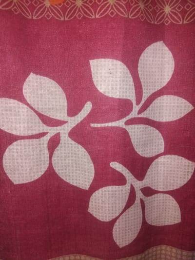 The image shows a close-up of a fabric with a maroon background and a pattern of white floral shapes. The fabric has a textured appearance due to the weave. At the top of the image, there is a border with a decorative pattern of interconnected diamond shapes and curved lines in a light beige or off-white color. The floral motifs are stylized and resemble leaves or petals arranged in a cluster. The lighting suggests the image was taken indoors, possibly in a home environment. There are no people, animals, or specific landmarks visible in the image, and therefore no information can be provided about the location in Haveli Jhangranwali, Pakistan.