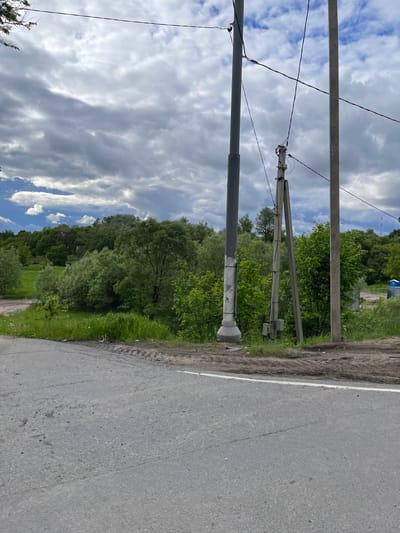 The image presents an oblique view of a roadside scene in Semenovskoe, Russia. On the left side, there are two utility poles, one with visible power lines, set against a backdrop of cloudy blue sky. The poles are adjacent to a strip of green vegetation, beyond which lies a dense line of trees. The right side of the image is dominated by a paved road with a white shoulder line, showing typical wear and tear. The perspective suggests the shot was taken from a low angle, with the camera angled upwards towards the sky. No people are visible in the scene.