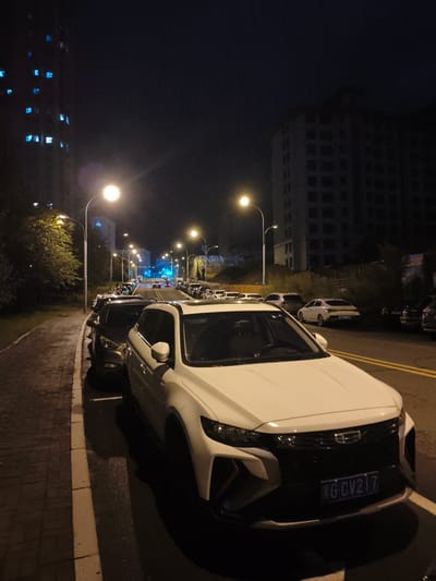 The image depicts a nighttime scene on a street in Jixi, China, likely in a residential or mixed-use area. A white SUV is parked on the right side of the road, facing away from the camera. The vehicle has a license plate that reads "G CV217". Several other cars are parked along the street, both in front of and behind the white SUV. The street is illuminated by streetlights. On the left side of the road, there is a sidewalk made of paving stones and a tree-lined area. In the background, there are multi-story buildings, one of which appears to be under construction or abandoned, with dark windows. Another taller building on the far left has several illuminated blue windows, suggesting it is occupied. The sky is dark, indicating it is nighttime. The ground appears wet in places, possibly from recent rain. There are no people visible in the image. The overall atmosphere is quiet and still, characteristic of a late hour in a city.