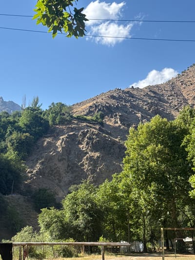 The image depicts a mountainous landscape with lush green trees in the foreground and a rocky, barren mountain face in the background. The sky is a clear blue with a few white clouds. In the lower part of the image, there is a wooden railing, and beyond that, some trees and what appears to be a small, possibly abandoned, structure. The overall scene suggests a natural, possibly rural or park-like, setting. While the image does not provide explicit visual cues to pinpoint the exact location within Мирата, Tajikistan, it aligns with the general topography and vegetation that might be found in mountainous regions of the country. The lighting suggests it is daytime, likely late morning or early afternoon, given the bright sun and clear sky. There are no people or discernible animals visible in the image. The presence of overhead wires, possibly for electricity or communication, hints at some level of human infrastructure in the vicinity, though it is not immediately intrusive.