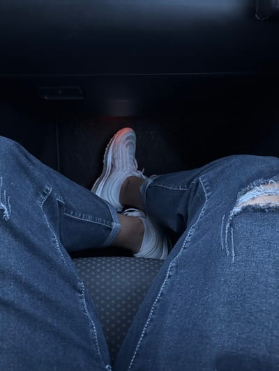 The scene depicts a first-person view of a person's lower body within the dark interior of a vehicle, located in Stara Reka, Bulgaria. The subject is wearing dark-wash denim jeans with visible distress and tearing on the right knee, and both pant legs are cuffed above the ankles. The feet are adorned with white athletic shoes, characterized by a wavy, multi-layered design, and are positioned with ankles crossed, resting on an unseen surface. The shoes appear to be Nike Air Max 97s. Between the legs, a dark grey upholstered surface with a pattern of small, light-colored dots is visible, consistent with a vehicle's seat fabric. The background above the feet is dark, possibly the lower dashboard or footwell area, with a subtle red light emanating from the lower right side and a small rectangular depression visible in the upper left.