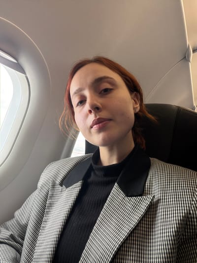 A female individual is depicted in a close-up shot, seated within the cabin of an aircraft. The location is on board an aircraft, likely at or near Sabiha Gökçen International Airport (SAW), which serves the Tuzla district of Istanbul, Turkey.

The subject is a young adult female with fair skin and reddish-brown hair, styled away from her face. She wears a small stud earring in her left earlobe. Her lips are lightly colored, and her gaze is directed towards the camera with a neutral expression.

She is dressed in a black ribbed turtleneck sweater, overlaid by a black and white small-checkered blazer with solid black lapels.

To her left, an oval-shaped airplane window is visible, revealing an overexposed external scene indicating daylight. The interior structure of the aircraft, including a portion of the ceiling and an overhead panel, is discernible. A dark-colored aircraft seatback is partially visible to her right. The image is captured from a slightly low angle, with the subject occupying the majority of the frame.