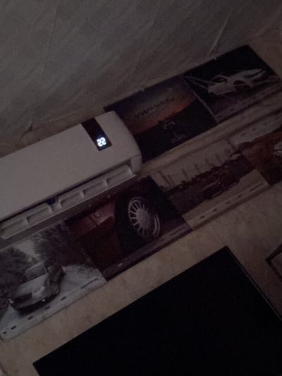 The image shows a tilted view of a wall. The wall is covered with wallpaper with photographic patterns and textures including a portrait, writing, a car wheel, and what appear to be architectural elements. An air conditioning unit with a digital display showing '22' is attached to the wall. A dark rectangular object is also visible on the wall. The specific location within the city of Varbak, Bulgaria, cannot be determined from the information contained in the image.