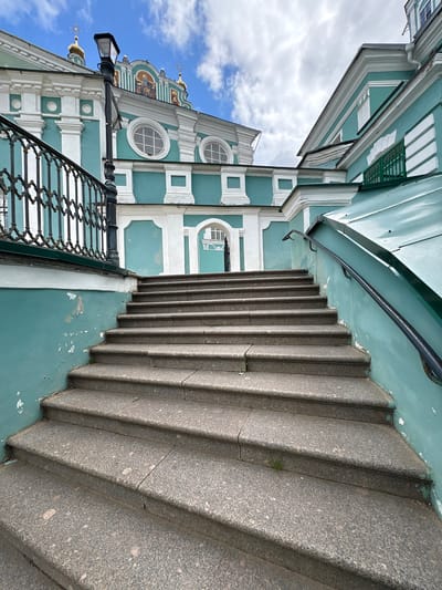 The image shows an outdoor view of a grand staircase leading up to a building with striking turquoise and white architecture. The staircase is made of rough-textured granite and has a black metal handrail on the right side. The building features ornate white detailing, round windows with intricate grilles, and gold-domed structures visible on the rooftop against a cloudy blue sky. A black street lamp stands to the left of the stairs, near an elaborate black metal fence. The turquoise walls of the building show signs of wear, with some peeling paint. The overall scene suggests a historical or religious site in Demenshchina, Russia. There are no people visible in the image.
