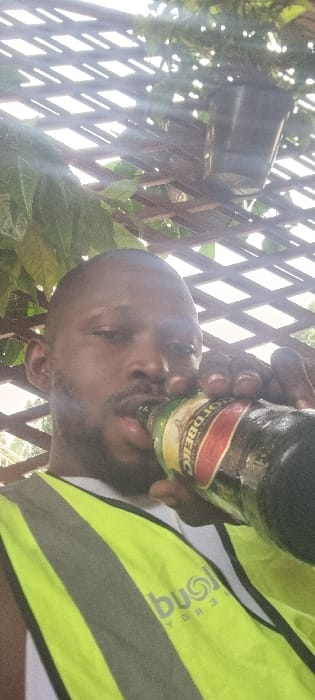 A man is depicted drinking from a dark bottle outdoors. His head is tilted back as he brings the bottle to his mouth. The bottle shows condensation droplets and features a label with red, yellow, and green accents. Text on the label appears to be in a Cyrillic-like script, possibly "СГОТОВЕКС". The man is wearing a bright yellow or lime green reflective safety vest with grey horizontal stripes. A partial logo and text, resembling "rông" and possibly "ENERGY" or "strong," are visible on the vest.

The background features a light-colored wooden lattice or trellis, interspersed with green foliage. Bright daylight illuminates the scene, with noticeable lens flares in the upper left, suggesting a sunny outdoor environment. The activity is the man drinking, likely taking a break. The setting is an outdoor space, possibly a garden or patio, located in Alagbado, Nigeria.