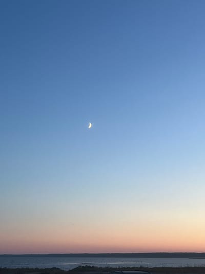 The image depicts a clear sky transitioning from a deep blue at the top to lighter hues of pink and orange near the horizon. A slender crescent moon is visible in the upper portion of the sky. Below the sky, a body of water stretches across the frame, with a faint line of land visible on the opposite shore. The foreground shows a dark, indistinct area that could be land or a structure, with a dark object that appears to be a vehicle partially visible at the bottom edge of the image. The scene suggests an outdoor setting, possibly during dusk or dawn.