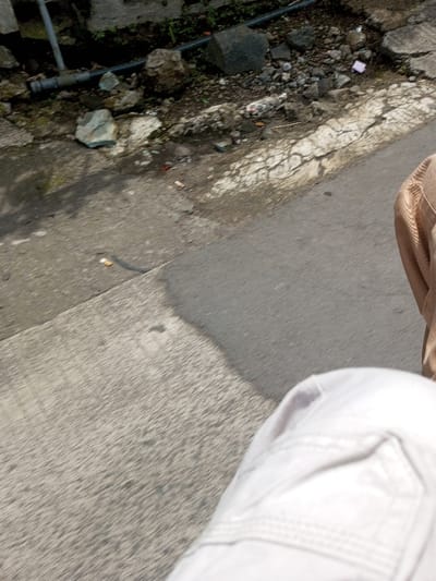 The image depicts a roadside environment in the city of Namthoung, Laos, viewed from an oblique, downward angle. In the lower right foreground, the upper thigh of an individual wearing light-colored, possibly white, cargo-style pants is visible, alongside a section of a tan or brown garment. The central and lower-left areas are dominated by a gray paved surface, likely asphalt or concrete, with a large, dark shadow extending diagonally across it from an unseen object to the left. The background features an unkempt embankment consisting of loose earth, various sized rocks, and sparse green vegetation. Several items of debris are present, including multiple white cigarette butts on the edge of the paved surface and scattered fragments of light-colored material, along with a small pink object on the embankment. Sections of a grey metal pipe and a dark, possibly plastic, pipe are visible horizontally along the upper left portion of the embankment. The paved surface where it meets the embankment exhibits visible cracking and weathering.