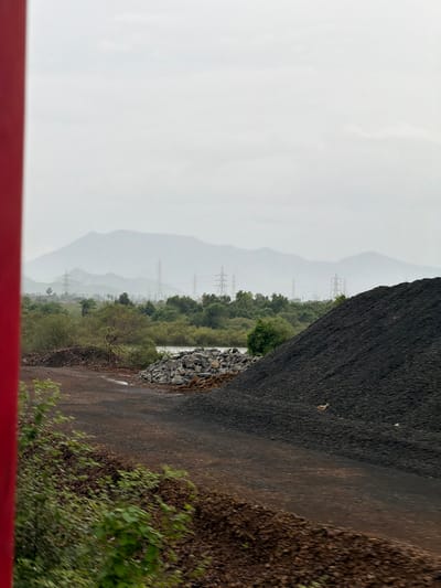 The image depicts an outdoor industrial scene in Saphale, India, with a focus on a large pile of black, granular material, likely coal or ore, dominating the right foreground. A dirt path or road runs through the middle ground, separating the large pile from a smaller pile of grey rocks and debris to its left. Further into the background, there is a body of water with green foliage and trees lining its banks. Beyond the vegetation, a hazy landscape of rolling hills or mountains is visible, punctuated by several tall electricity pylons with visible transmission lines. The sky is overcast and appears to be a pale, muted grey. A vertical red pole is partially visible on the extreme left edge of the frame, suggesting the perspective might be from a moving vehicle or a structure. The overall impression is one of an industrial area situated in a natural, somewhat undeveloped environment.