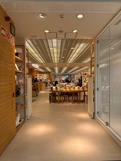 The image shows the interior of a shop in Ondara, Spain. The space features a long corridor lined with shelves filled with various products. Further back, there's a table with chairs, suggesting a seating or display area. A couple of people are present, seemingly browsing the merchandise. The foreground reveals a wooden counter with display cases below, holding items such as boxes, lamps, and what appears to be a Prada product. The overall environment is well-lit, with a clean and modern design. The floor is tiled, and the walls are a combination of plain and textured surfaces.