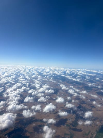 The image is an aerial view of the city of Raczkowa, Poland, as seen from an airplane. The scene is dominated by a vast expanse of white, puffy cumulus clouds scattered across a clear blue sky. Below the clouds, the landscape of Raczkowa is visible, characterized by a patchwork of brown and green fields, indicating agricultural activity. Several roads and what appear to be clusters of buildings, suggesting urban or semi-urban areas, can be discerned through the breaks in the clouds. The sun is casting shadows from the clouds onto the ground, creating a dappled light effect. No people or specific actions are discernible from this high-altitude perspective.