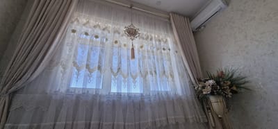 The image depicts an interior scene focused on a window covered by ornate, sheer curtains. The curtains have a lace-like pattern with floral motifs and a layered hem. A decorative Islamic calligraphy piece, possibly spelling "Allah," hangs from a cord in the center of the window, with a small tassel below it. To the right of the window, a cream-colored air conditioning unit is mounted high on a textured, light-colored wall. On the right side of the image, a tall vase with an arrangement of dried flowers and greenery stands against the same textured wall. The room appears to be a living space or bedroom, with heavy drapes on either side of the window. The lighting suggests it is daytime, with light filtering through the sheer curtains. The location is identified as Balkanabat City, Turkmenistan.