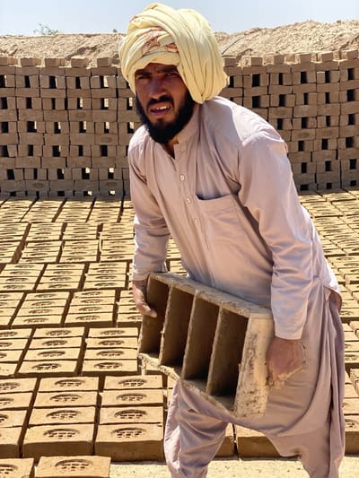 A man in Deh-E Bagh, Afghanistan, wearing a turban and traditional clothing, carries a mold for bricks. He is surrounded by rows of freshly made bricks, with a stacked brick wall in the background.