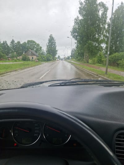 This is a photograph taken from inside a vehicle, looking out through a rain-streaked windshield. The view captures a wet road with visible puddles, suggesting recent or ongoing precipitation. Trees and buildings line the road, with one house clearly visible on the left side of the frame, appearing somewhat aged and weathered. The sky is overcast and grey. The dashboard of the car is in the foreground, showing a speedometer indicating a speed between 0 and 20 km/h, and an odometer reading 345853. The time displayed is 2:03. Streetlights are visible on the roadside. The overall impression is that of a quiet, overcast day in a rural or semi-rural area of Viļāni, Latvia, with the driver experiencing inclement weather.
