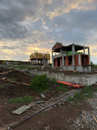 The image shows a construction site of a multi-story building in Cherni Vrah, Bulgaria. The building's concrete frame is partially complete, with brick infill on some levels. Construction materials are visible, including lumber, concrete, and rebar. Orange pipes are laid along the ground near the structure. The surrounding area is undeveloped, with soil, grass, and scattered gravel. The sky is cloudy with diffused sunlight.