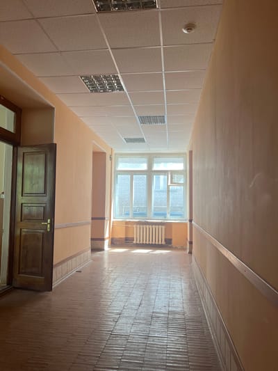 The image depicts a sunlit, empty corridor in a building. The corridor has pale orange walls with a light-colored skirting board and a horizontal silver trim running along the walls. The ceiling is a suspended white tile system with integrated fluorescent light fixtures and a smoke detector. The floor is made of narrow wooden planks laid horizontally, showing signs of wear.
On the left side of the corridor, an open, dark wooden door with a brass handle is visible. Further down the corridor, a large window with multiple panes allows natural light to flood in, casting shadows on the floor and illuminating a white radiator beneath it. The window overlooks an exterior with visible brickwork. The overall impression is of a utilitarian, perhaps institutional, interior space in daylight. The location is identified as Караваево, Russia. There are no people or animals present in the image.