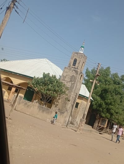 The image depicts a daytime scene on an unpaved road in Sumaila, Nigeria, with a mosque dominating the center-left. The mosque features light yellow-tan walls and a steeply pitched, white corrugated metal roof. A prominent, weathered, square-based minaret, constructed of mottled grey-tan concrete or stucco, stands adjacent to the main mosque structure. The minaret has arched window openings near its summit and a smaller rectangular opening below, topped with a white structure and a green dome/finial. A low, similarly weathered wall separates the mosque complex from the dirt road, behind which a leafy green tree is planted.
A young child, dressed in a light blue long-sleeved shirt and light trousers, walks on the dirt road towards the right, positioned near the low wall. Further to the right, two adult males walk away from the camera: one in a white t-shirt and light pants, and the other in a pink collared shirt and dark pants. Behind these individuals, partially obscured by a tree, a wooden stand or kiosk is visible. Overhead, utility poles with multiple black wires traverse the clear blue sky. The ground throughout the scene is composed of dry, light-colored dirt.