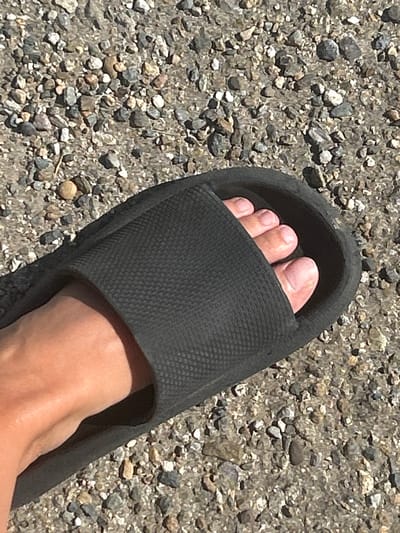 A left human foot is partially visible, inserted into a black slide sandal. The sandal is resting on a surface covered with small, grey and brown pebbles and gravel. The foot shows four toes and part of the sole. The background consists of more of the same pebbly ground. The lighting suggests an outdoor environment, likely during daylight hours. There are no other people or objects clearly discernible in the image besides the foot, sandal, and ground. The image does not contain enough information to determine a specific location within Temryuk, Russia.