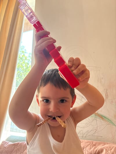 The image displays a young child indoors in Barakovo, Bulgaria. The child, appearing male with short, dark brown hair and dark eyes, wears a white sleeveless undershirt and looks directly at the camera with an open-mouthed smile. Two pretzel sticks, one visibly covered in sesame seeds, are held horizontally in the child's mouth. The child holds a red and clear modular plastic object or toy overhead with both hands; the left hand grasps a red segment near the clear portion, and the right hand holds a lower red segment. The background features a light-colored wall extensively covered with scribbles and drawings in multiple colors (e.g., black, gray, red, green, orange). To the left, a window provides natural light, framed by light-colored curtains, and through it, sunlit green foliage is visible. A quilted pink fabric surface is present in the lower right and left portions of the frame.