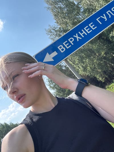 A close-up, rotated selfie shows a person's face and upper body against a backdrop of trees and a bright blue sky with scattered clouds. The person has blonde hair, fair skin, and is wearing a black ribbed top. They are touching their face with their right hand, which is adorned with two rings. A smartwatch is visible on their left wrist. To the left of the person, a blue directional sign with white text is partially visible, indicating a direction with a white arrow. The text on the sign reads "ВЕРХНЕЕ ГУЛЯЕВО" which translates to "Verkhnee Gulyaevo". The lighting suggests it is daytime, likely sunny, with dappled sunlight filtering through the trees. The overall mood appears casual and relaxed.