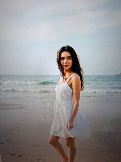 A medium shot captures a young woman with long, dark, wavy hair standing on a sandy beach. She is wearing a white, sleeveless sundress. The woman is looking to her left, with her body angled slightly towards the camera. Behind her, the ocean stretches out to the horizon, with gentle waves lapping at the shore. The sky above is a pale, hazy blue, with a few indistinct shapes that might be birds or distant objects on the water. The overall lighting suggests it is either early morning or late afternoon, with a soft, diffused light. The image is a photograph, and the foreground shows a sandy beach with some faint footprints and a piece of dried grass.