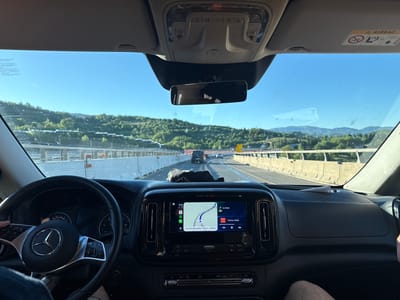 The image captures the interior of a vehicle from the driver's perspective, showing a person's hand on the Mercedes-Benz steering wheel and a passenger's leg on the right. The car is traveling on a multi-lane highway or motorway, characterized by concrete guardrails and distant green, tree-covered mountains under a clear blue sky. Other vehicles, including trucks, are visible further down the road.

The infotainment system on the dashboard is active, displaying navigation and music. The time is 18:48, with an estimated arrival at 20:39 after 1 hour and 51 minutes, covering 200 km. The navigation indicates a turn in 2.4 km, and the language "Arrivo previsto" (Estimated arrival) confirms an Italian setting, aligning with Barberino di Mugello. Music titled "Famam (Edit)" is playing. An overhead console with controls and a rearview mirror are seen above, while a sun visor on the right displays an "AIRBAG" warning. The windshield appears speckled with dirt or dust. Partially visible stickers on the left side of the windshield include text in Portuguese such as "PARA A RODAGEM". The speedometer, visible through the steering wheel, shows markings for speeds up to 160 km/h. The scene depicts a calm, sunny late afternoon road trip through a scenic, mountainous region.
