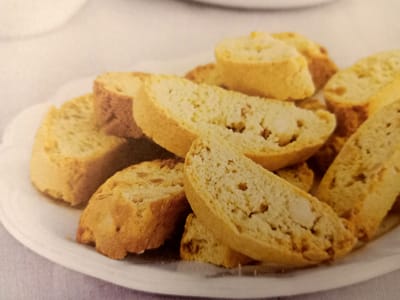 A white plate filled with sliced biscotti is shown on a white tablecloth. The biscotti are golden brown and appear to have pieces of nuts embedded within them. The plate is partially visible, as is a portion of the tablecloth. The image is a close-up shot of the biscotti. There is no indication of the city of Rumaithiya, Kuwait in the image.