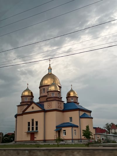 The image captures an Orthodox church located in Velyki Hai, Ukraine, under an heavily overcast, grey sky. The church is a multi-domed structure, featuring five prominent golden domes, each surmounted by an Orthodox cross. The largest dome is central, with two smaller domes on either side. The roofs of the church are a distinct blue color, contrasting with the light beige exterior walls. Geometric decorative patterns, in a darker brown or red hue, are visible on the upper sections of the beige walls, particularly beneath the domes. The building exhibits multiple rectangular windows with white frames; a series of three tall, narrow windows are present above a red entrance door on the front facade. A smaller, blue-roofed porch is visible on the right side of the structure. Numerous black power lines span horizontally across the upper portion of the frame, partially intersecting the sky and the top of the central dome. The foreground consists of a blurred horizontal barrier, possibly a wall, above a strip of green grass. In the background, beyond the church, several smaller buildings with red roofs and sparse trees are discernible. No individuals are present in the scene.