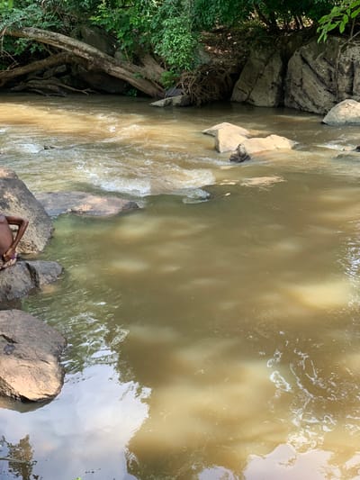 The image shows a body of water with muddy brown water, likely a river or stream. The water is moving swiftly and is slightly choppy. There are several large rocks in and around the water. One large rock in the foreground has a young person sitting on it, facing the water. The person is wearing a dark shirt and a dark skirt. The scene is in a wooded area with thick vegetation on the far bank. The scene is located at the coordinates 7.119 latitude, 5.362 longitude, in the city of Egbeda, Nigeria.