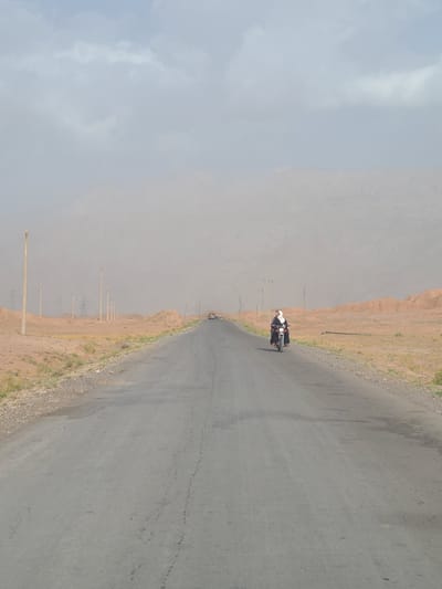 This media file depicts a scenic view of a road in a dusty, arid environment, likely Bostan, Pakistan. The focal point is a motorcycle with two riders, dressed in casual attire, traveling along the asphalt road. The road stretches into the distance, bordered by a dry, sandy terrain with sparse vegetation. In the background, imposing, dusty hills rise up, adding to the rugged landscape.

The scene appears to be set during the day, with a hazy sky suggesting either dust or a lightly overcast condition. The presence of utility poles and wires along the side of the road indicates that this is likely a route that serves a populated area or is part of infrastructure development. The overall atmosphere is one of travel and exploration through a remote and natural landscape. There is no visible text in the image.