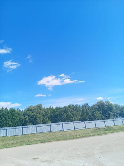 The image captures a bright, sunny day with a clear blue sky dotted with scattered white clouds. In the foreground, a dirt road runs diagonally across the bottom of the frame. Beyond the road, a patch of green grass slopes upwards towards a long, light blue metal fence. This fence appears to be made of vertical corrugated panels supported by posts. Behind the fence, a dense line of green trees forms a natural barrier, with the foliage indicating a summer season. The scene is tranquil and appears to be located in an open area, possibly on the outskirts of a populated area, consistent with the rural setting of Пралетарскі сельскі Савет, Belarus. There are no people or animals visible in the image, and no objects of significant interest are present beyond the natural landscape and the man-made fence and road. The overall impression is one of a peaceful, natural environment bathed in sunlight.