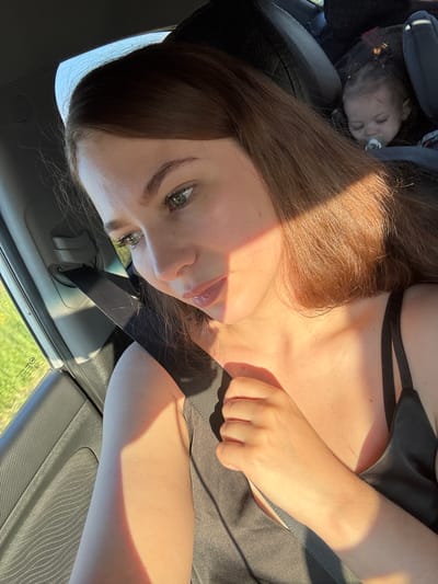In Kvarsa, Russia, a woman with long, auburn hair, wearing a black tank top, sits in the passenger seat of a car. Sunlight streams through the window, casting a bright diagonal shadow across her face and chest. Her gaze is directed downward and to the side. In the background, a baby is visible in a car seat, sleeping with a pacifier in its mouth. The car's interior, including a seatbelt and door panel, is also visible. Outside the car window, a field of green foliage can be seen.