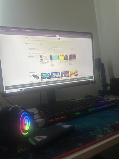The image shows a desk with a computer monitor, keyboard, and mouse. The computer screen displays the Fiverr website, which is an online marketplace for freelance services. The user's name "kristian780" is visible on the screen, indicating a personalized welcome. Various categories of services are listed, such as Graphics & Design, Programming & Tech, and Digital Marketing. Below the welcome message, there are sections for "Recommended for you" and "Based on what you might be looking for," showcasing different gig offerings. A banner at the bottom of the screen informs the user about the website's use of cookies. The environment appears to be indoors, likely a home office or study area, with a wall visible in the background. The lighting suggests it might be evening or a dimly lit room. The presence of a keyboard with RGB lighting and a gaming mouse indicates a modern setup. There are no people visible in the image. The location of Madara, Bulgaria is not indicated by any visual cues in the image.